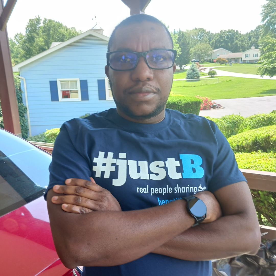 Man wearing glasses and a #justB shirt, standing with crossed arms on a porch, with houses and greenery behind.