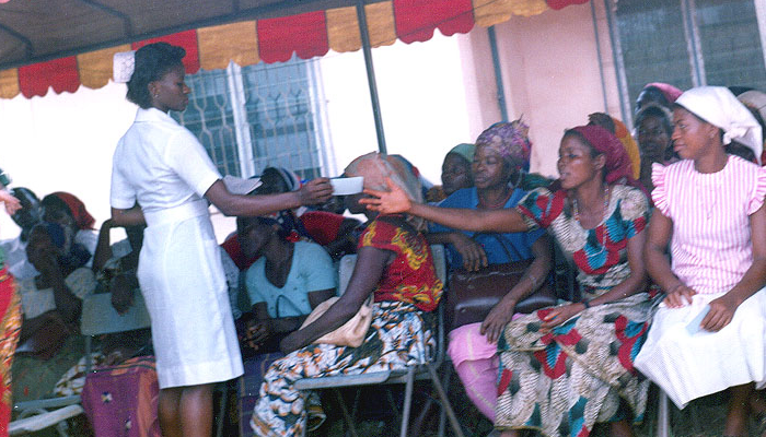 Nurse distributing paper slips to seated women in an outdoor clinic setting.