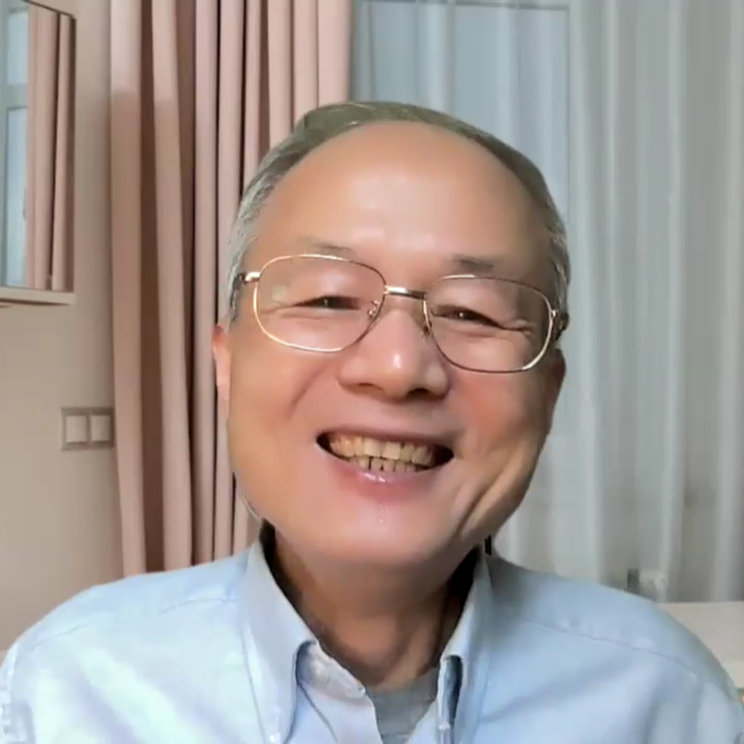 Older man with glasses smiling warmly, wearing a light blue shirt, and standing in a softly lit room.