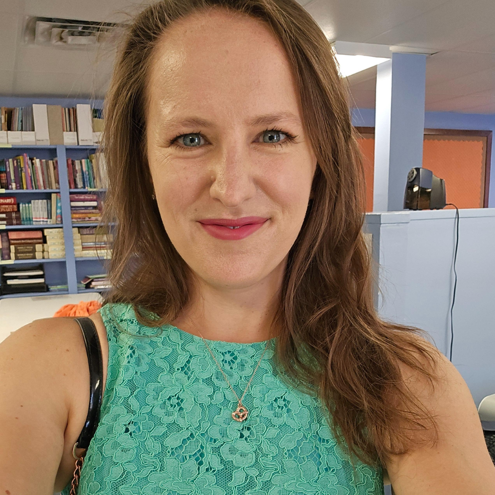 A woman in a turquoise lace top smiles while taking a selfie in a room with bookshelves in the background.