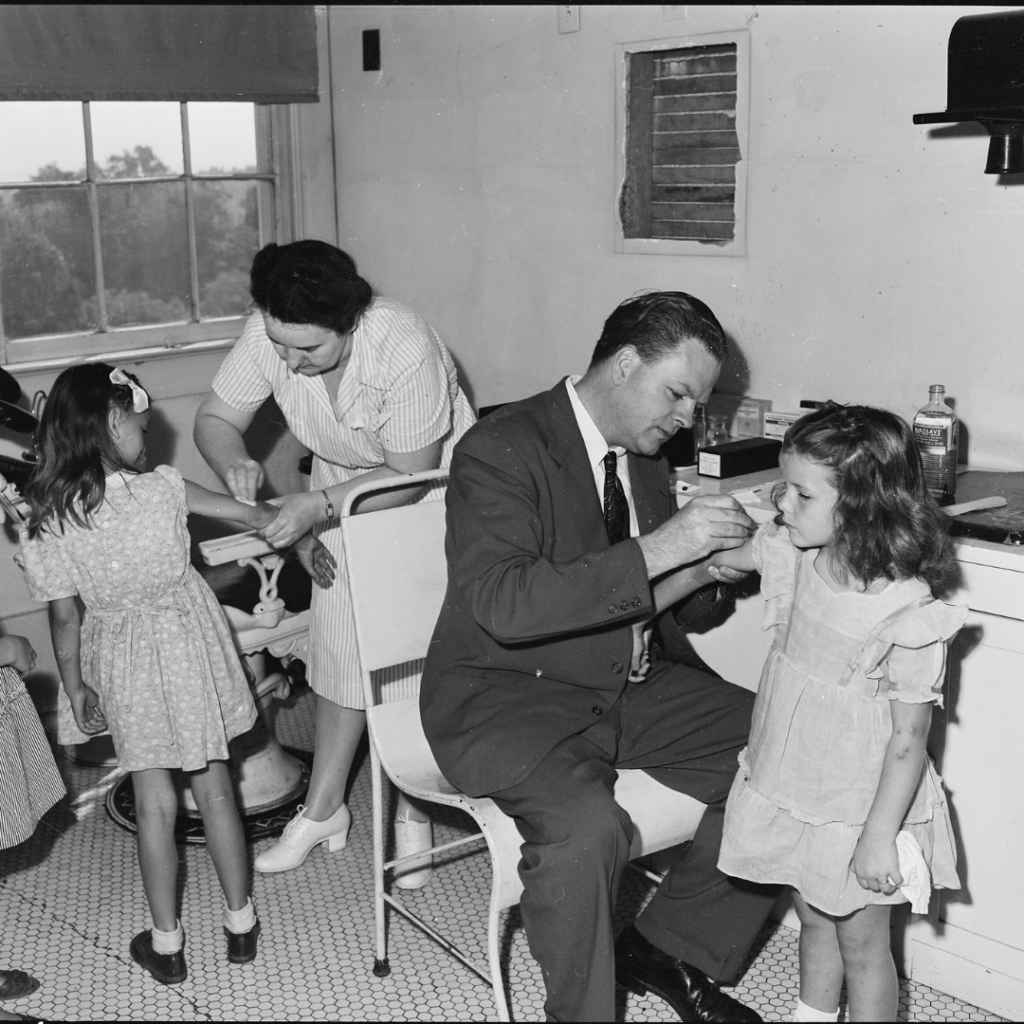 A black and white photo from the 1940s of two children receiving smallpox vaccines.